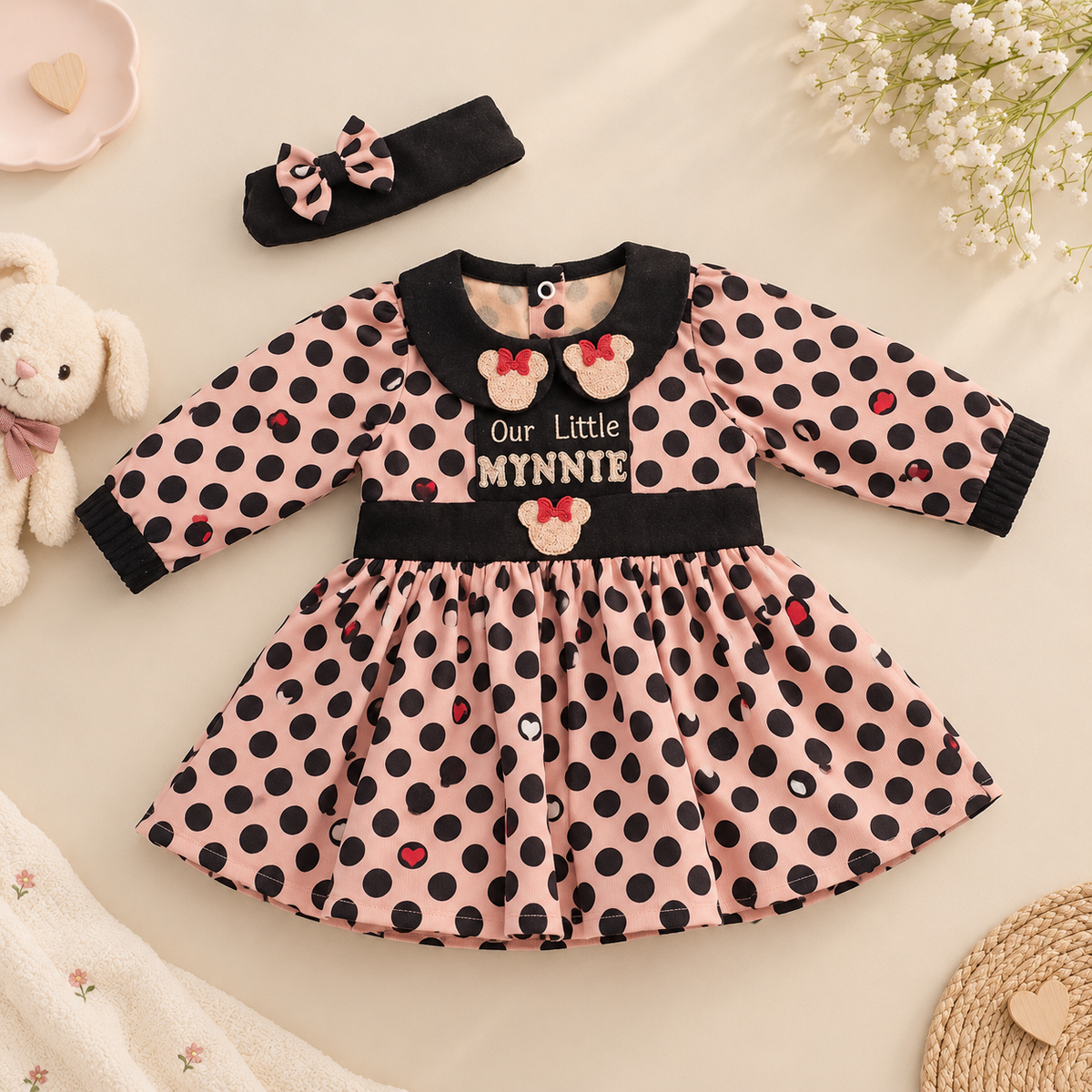 A baby girl's long-sleeve dress with a pink and black polka dot pattern, featuring a black velvet collar and cuffs, a matching black waistband, and Minnie Mouse-inspired embroidered details. A matching black and pink polka dot headband is placed above the dress on a beige fabric background with baby props including a plush bunny, flowers, and a woven mat.