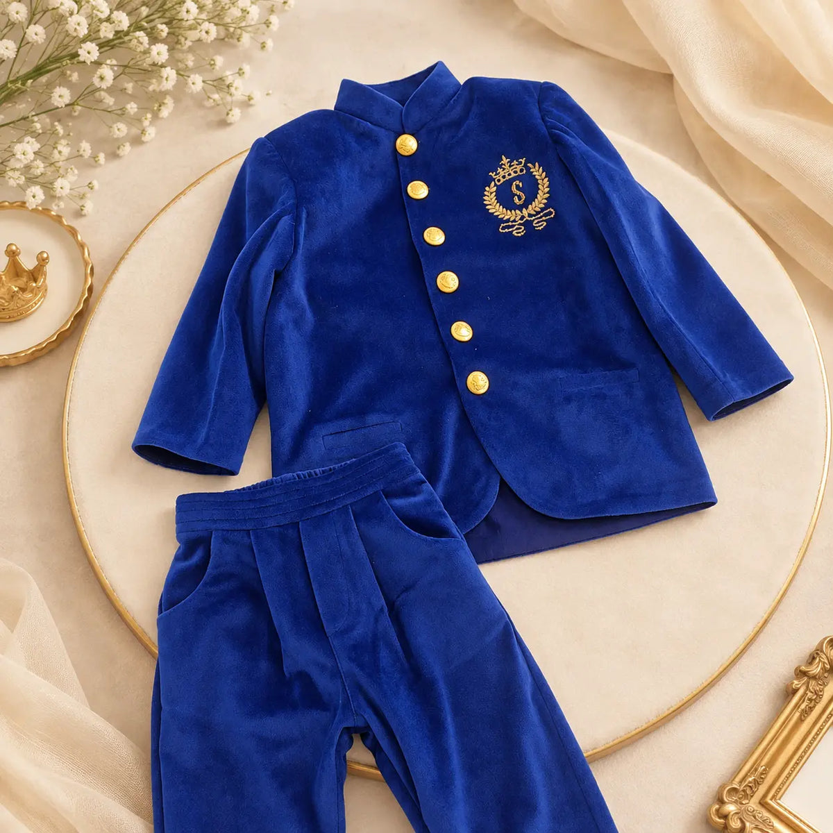 A royal blue velvet children's outfit set consisting of a long-sleeve jacket with gold buttons and a gold crest emblem, paired with matching pants, styled on a round beige platform with decorative dried flowers and fabric.
