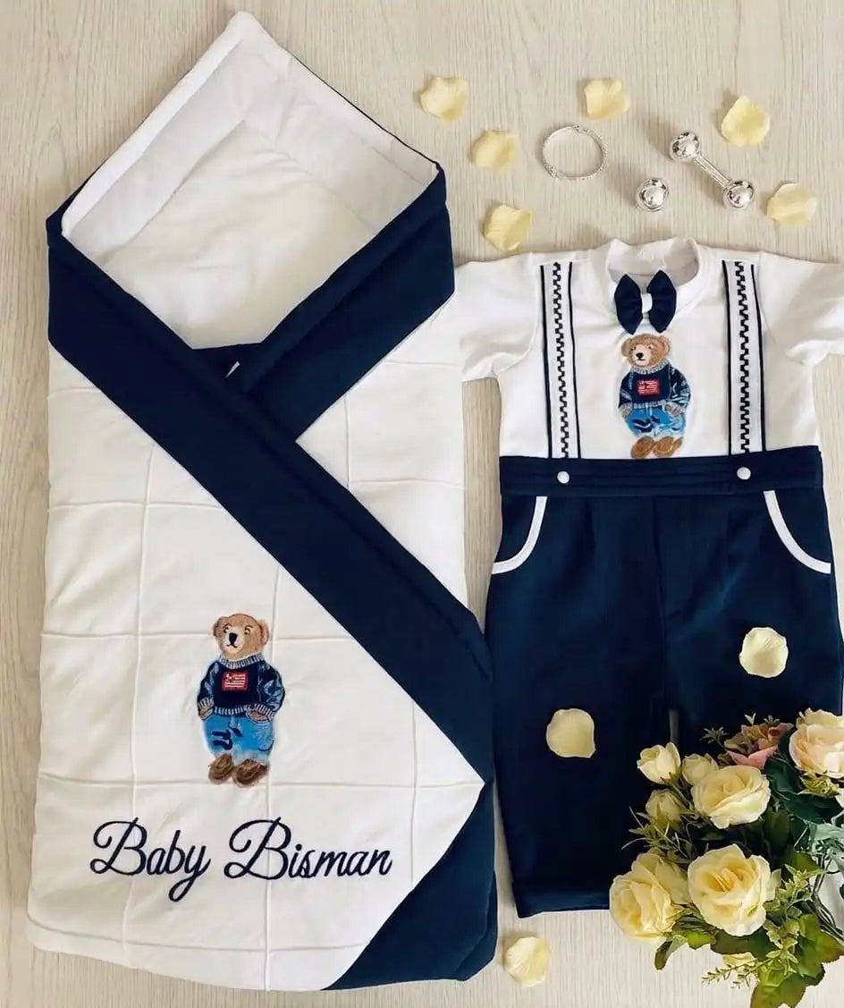A baby outfit and matching swaddle set laid flat on a light wood surface. The set includes a white long-sleeve romper with navy shorts, navy suspenders, a navy bow tie, and a teddy bear patch, plus a matching white and navy swaddle wrap with an embroidered teddy bear and the text 'Baby Bisman.