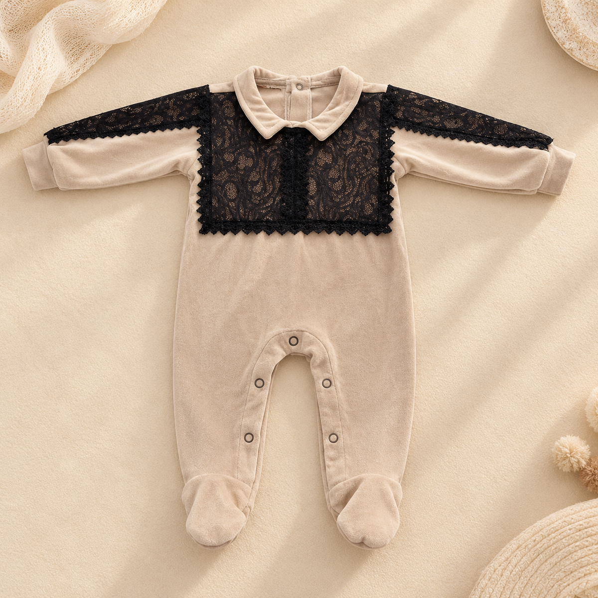 A baby footed sleeper in cream-colored velvet with a black lace yoke panel, laid flat on a soft beige fabric background with decorative props.