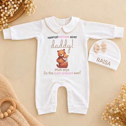 A white long-sleeve baby romper with a cute teddy bear graphic and birthday text, displayed alongside a matching white baby hat with a bow and the name 'RAISA' on a beige background with decorative dried flowers.