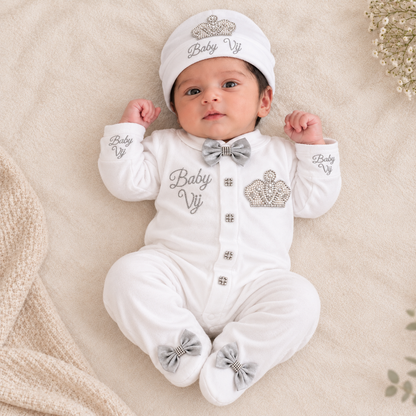 A baby lying on a soft cream blanket wearing a white footed onesie with silver-gray bow tie, silver crown embroidery, and matching beanie hat.