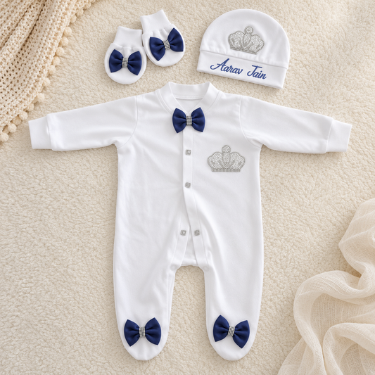 White baby footed sleeper with silver crown embroidery, navy blue bow tie at the neck, and navy blue bow ties on the feet, shown with matching booties and a beanie on a cream textured blanket.
