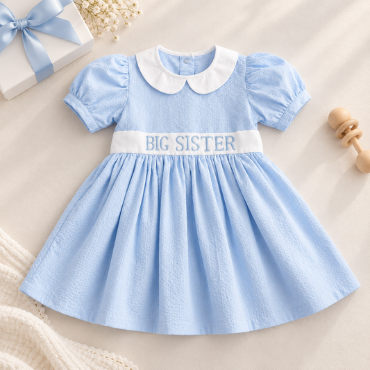 A baby girl's light blue dress with a white Peter Pan collar and a white waistband embroidered with 'BIG SISTER' in blue. The dress has short puff sleeves and is styled in a flat-lay on a beige background with a gift box, dried flowers, and a wooden rattle.