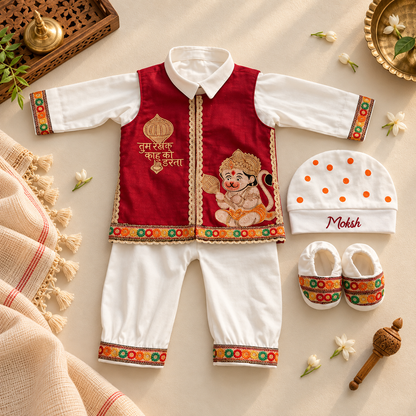 A baby clothing set laid flat on a warm beige surface, including a red and white traditional outfit with embroidered details, a white hat with orange dots and the name 'Mohsh', and matching white booties with red and multicolor trim. Decorative props like flowers, a brass pot, and woven textiles surround the set.