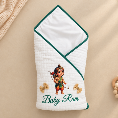 A white quilted baby swaddle blanket with dark green piping, featuring an embroidered illustration of Lord Ram and the text 'Baby Ram' in green script. The swaddle is displayed flat on a beige fabric background with a textured cream knit blanket and wooden beads as props.