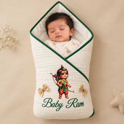 A sleeping newborn baby wrapped in a quilted cream swaddle wrap with green trim, featuring an embroidered illustration of Lord Krishna and the text 'Baby Ram'.