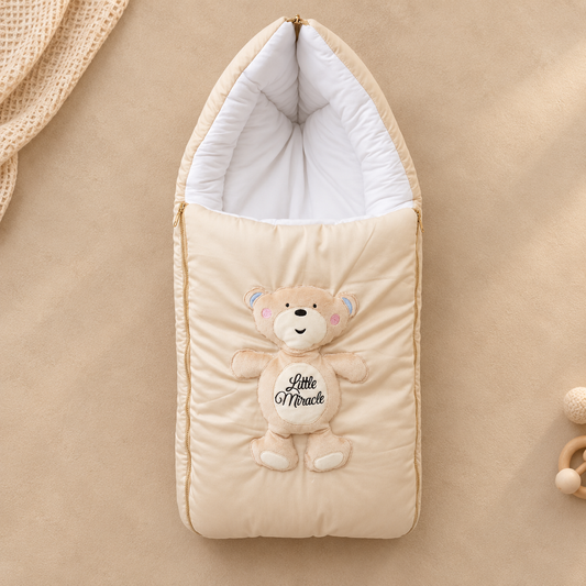 A cream-colored quilted baby footmuff with a plush teddy bear applique on the front. The bear has embroidered pink cheeks, blue ears, and a belly patch that reads 'Little Miracle'.
