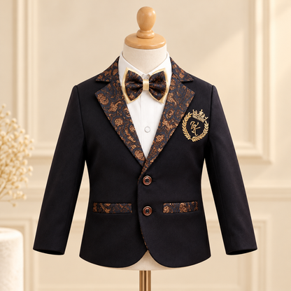 A boy's formal suit jacket displayed on a mannequin with a white dress shirt and matching bow tie. The jacket is black with ornate gold and brown patterned lapels, pocket flaps, and a gold crest on the chest.