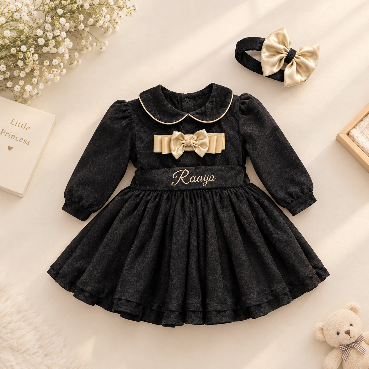 A baby girl's black dress with a gold bow and collar, personalized with the name 'Raaya' on the waist. The dress is styled in a flat-lay with a matching gold bow headband, baby flowers, a book, and a teddy bear on a cream background.