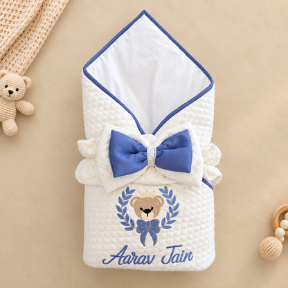 A white quilted baby swaddle blanket with blue trim, a large blue bow, and an embroidered teddy bear with the name 'Aarav Jain' below it, shown on a beige background with small toy props.