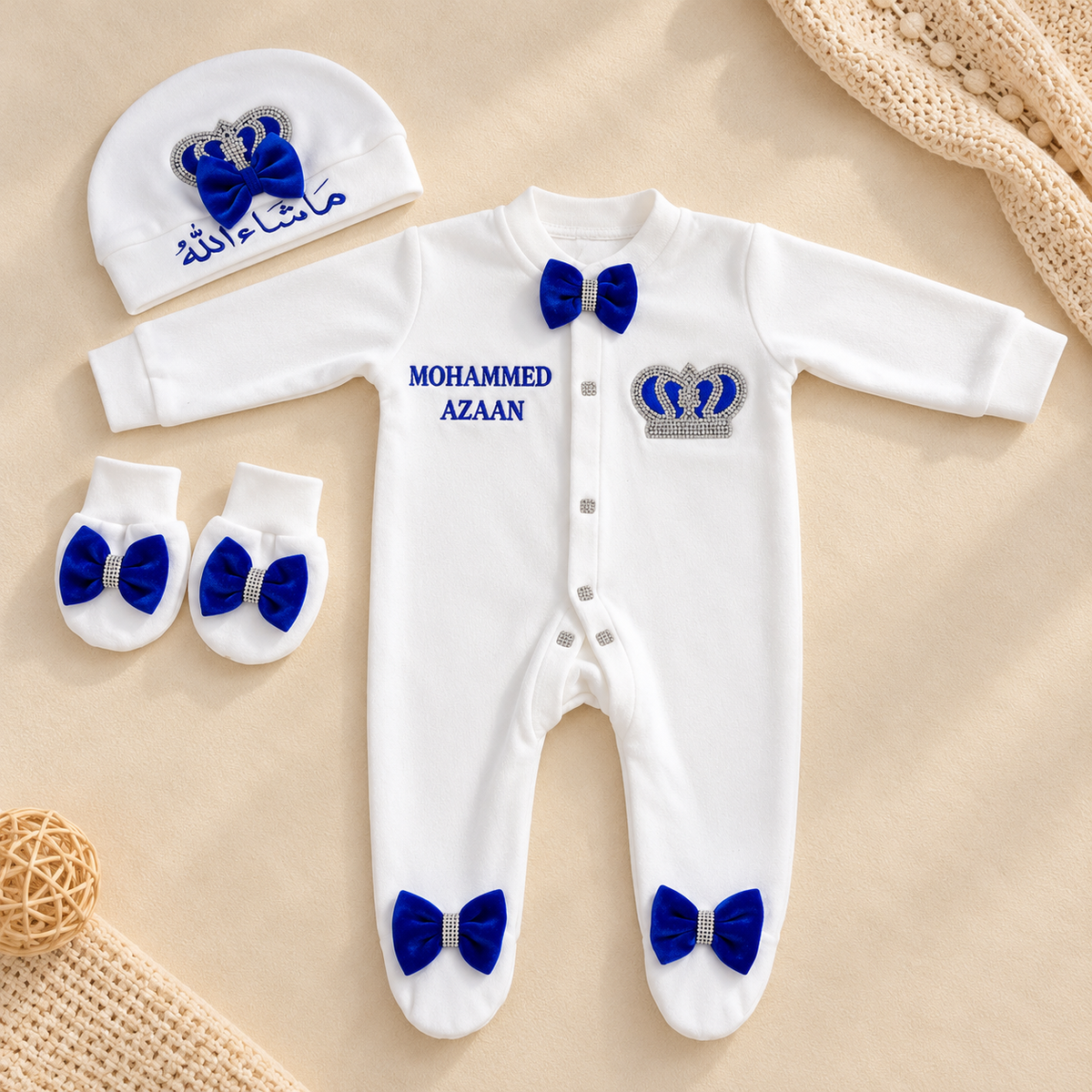 White baby footed sleeper set with royal blue bow accents, a matching beanie, and matching booties, laid flat on a beige background with decorative woven props. The sleeper has blue text reading 'MOHAMMED AZAAN' and a silver-and-blue crown motif on the chest.