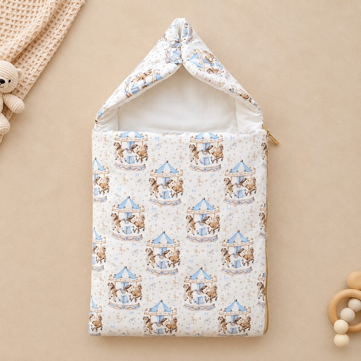 A baby sleep sack with a hood, featuring a repeating pattern of teddy bears on carousel rides in light blue and brown on a cream background. The sleep sack is displayed flat on a warm beige background with styled baby props, including a knitted blanket, wooden beads, and a wooden teether.