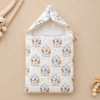 A baby sleep sack with a hood, featuring a repeating pattern of teddy bears on carousel rides in light blue and brown on a cream background. The sleep sack is displayed flat on a warm beige background with styled baby props, including a knitted blanket, wooden beads, and a wooden teether.