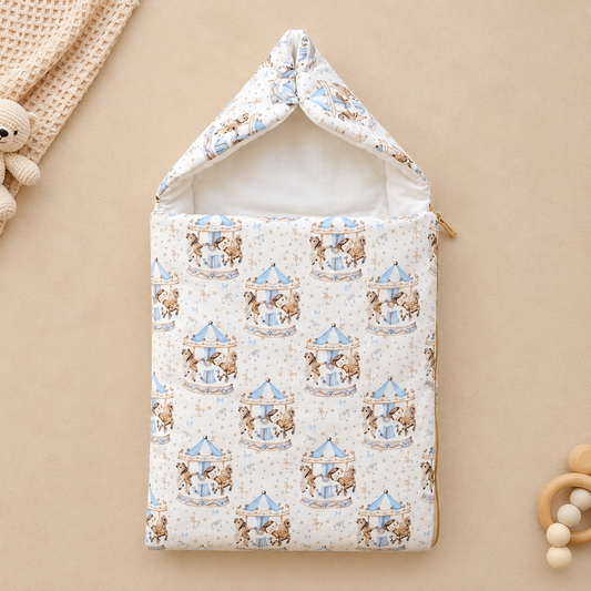 A baby sleep sack with a hood, featuring a repeating pattern of teddy bears on carousel rides in light blue and brown on a cream background. The sleep sack is displayed flat on a warm beige background with styled baby props, including a knitted blanket, wooden beads, and a wooden teether.