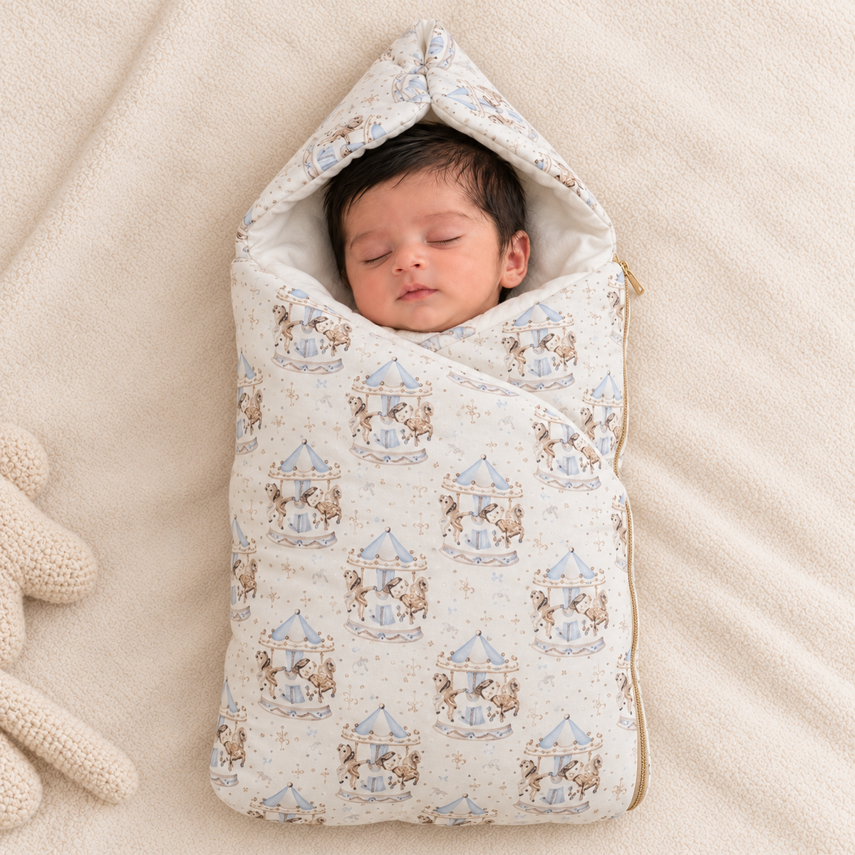 A sleeping newborn baby wrapped in a cream-colored hooded swaddle blanket with a blue and brown carousel pattern, lying on a soft beige blanket next to a knitted toy.