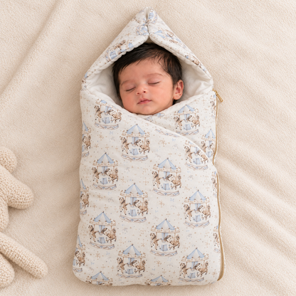 A sleeping newborn baby wrapped in a cream-colored hooded swaddle blanket with a blue and brown carousel pattern, lying on a soft beige blanket next to a knitted toy.