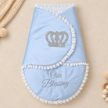 A light blue baby blanket or swaddle with white floral lace trim, featuring an embroidered silver crown on the upper fold and the words 'Our Blessing' on the lower section. The blanket is photographed flat on a warm beige background with soft decorative props.