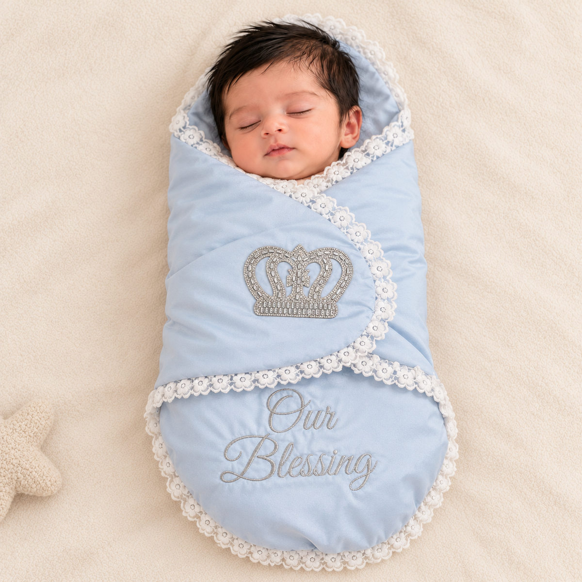 A sleeping newborn baby swaddled in a light blue blanket with white lace trim, featuring an embroidered silver crown and the words 'Our Blessing' on the front, lying on a soft cream-colored blanket.