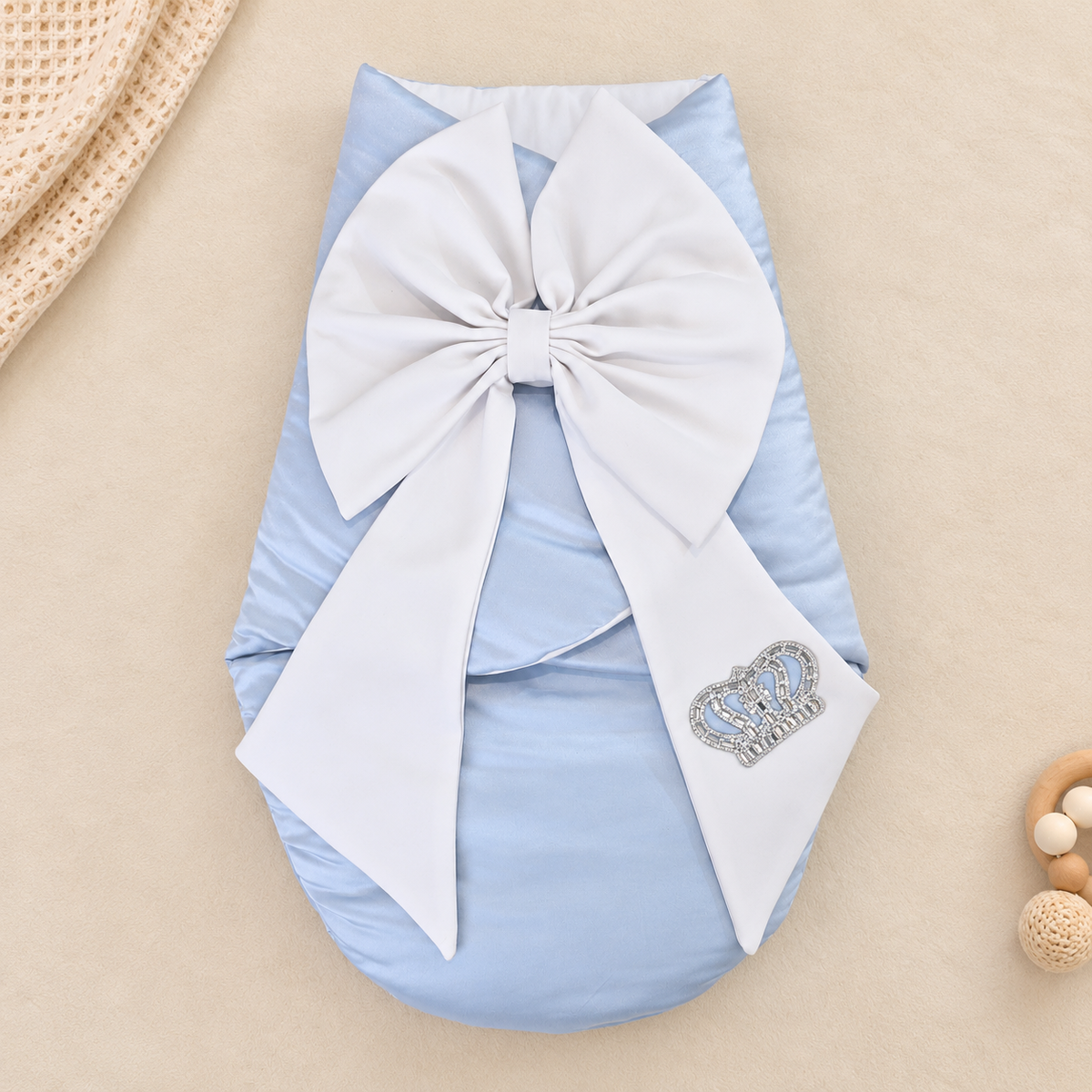A light blue baby swaddle wrap with a large white bow and a small rhinestone crown patch, laid flat on a beige background with styled props.