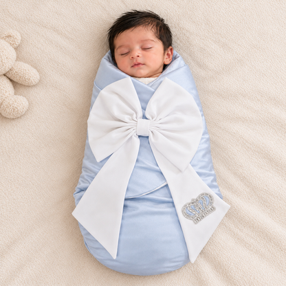 A sleeping newborn baby swaddled in a light blue wrap with a large white bow and a small rhinestone crown patch, lying on a beige blanket with a plush toy partially visible at the left edge.