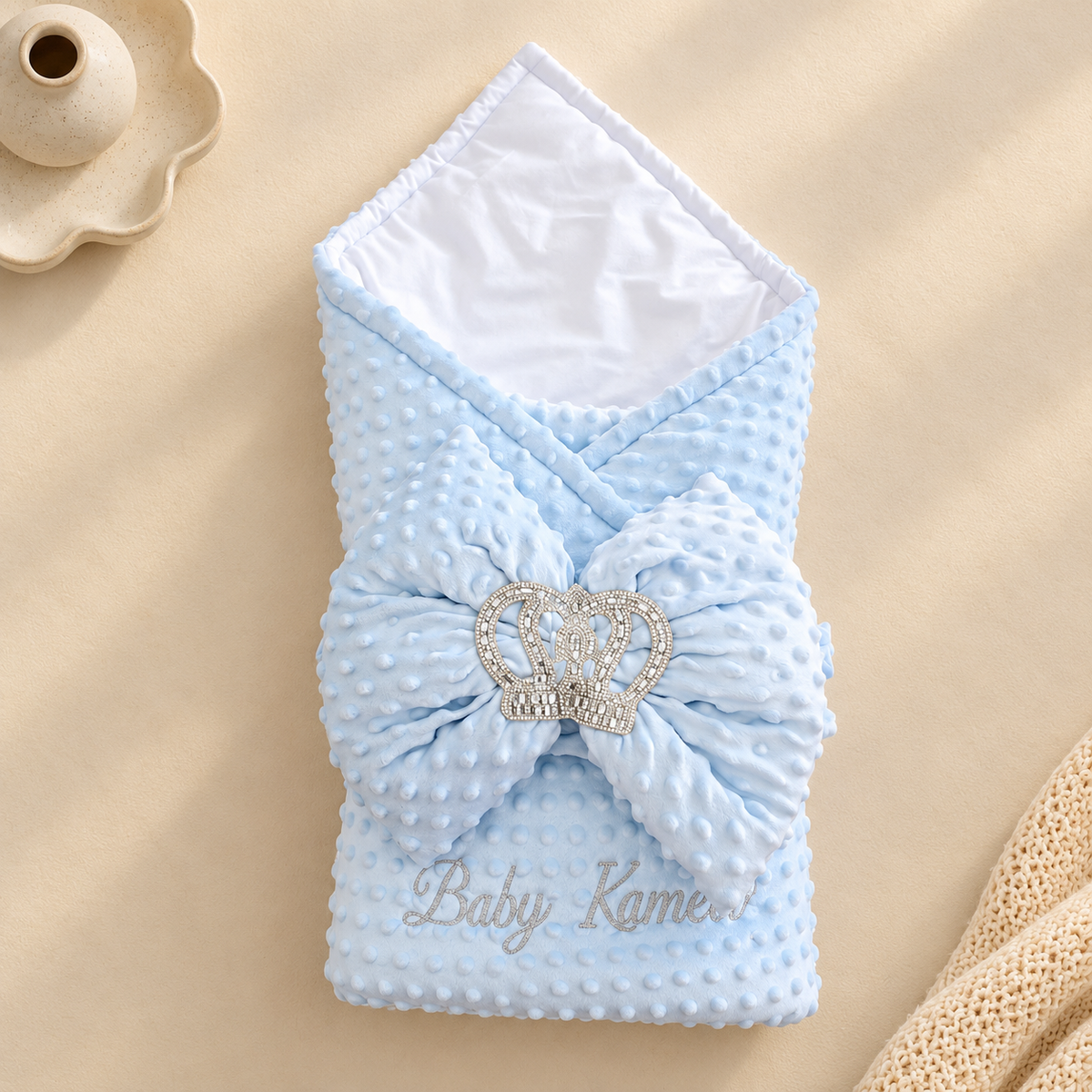 A light blue textured baby swaddle blanket with a large matching bow and a rhinestone crown embellishment, laid flat on a beige background with a scalloped dish and cream knit fabric as props.