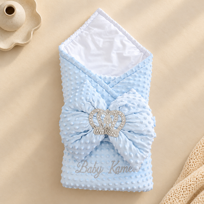 A light blue textured baby swaddle blanket with a large matching bow and a rhinestone crown embellishment, laid flat on a beige background with a scalloped dish and cream knit fabric as props.