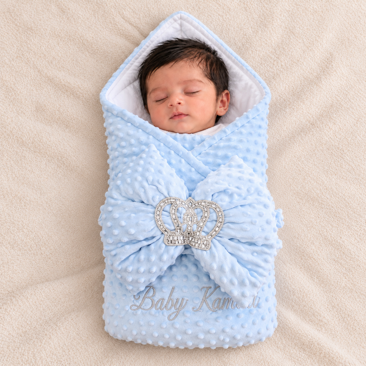 A sleeping newborn baby wrapped in a light blue dotted minky dot swaddle blanket with a large bow and a rhinestone crown embellishment, lying on a beige textured blanket. The brand name 'Baby Kamel' is embroidered in silver on the bottom right.