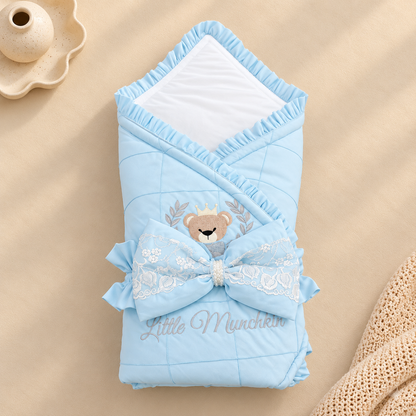 A light blue quilted baby swaddle blanket with a white inner lining, decorated with a large lace bow, a teddy bear face motif, and the text 'Little Munchkin'. The blanket is styled on a beige fabric background with a small ceramic vase and a knit textile prop.