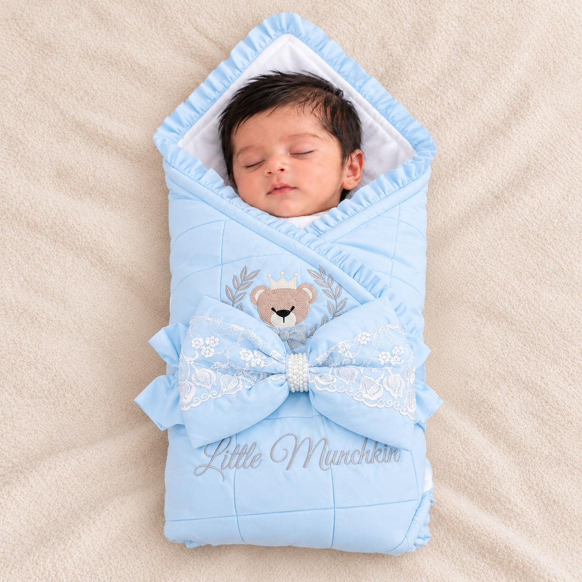A sleeping newborn baby wrapped in a light blue quilted swaddle blanket with a large bow, lace trim, embroidered teddy bear, and the text 'Little Munchkin' on a beige blanket.
