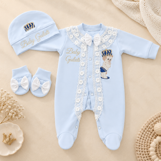 A baby footed sleeper set in light blue, including a long-sleeve one-piece with lace trim, a matching bonnet, and matching booties, arranged on a beige fabric background with decorative props.