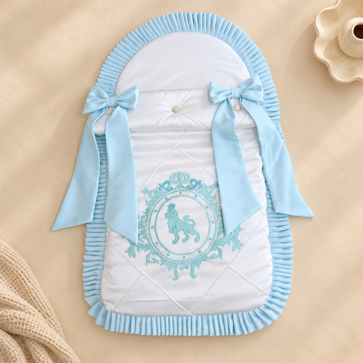 A light blue and white quilted baby bassinet insert with a lion emblem and ribbon bows, styled on a beige background with a knit blanket and ceramic cup.