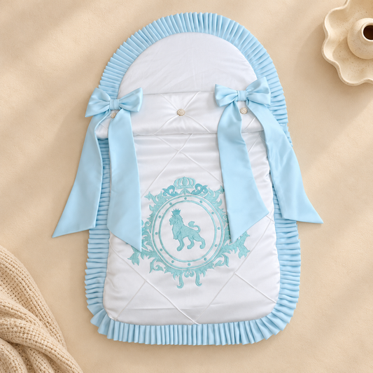 A light blue and white quilted baby bassinet insert with a lion emblem and ribbon bows, styled on a beige background with a knit blanket and ceramic cup.