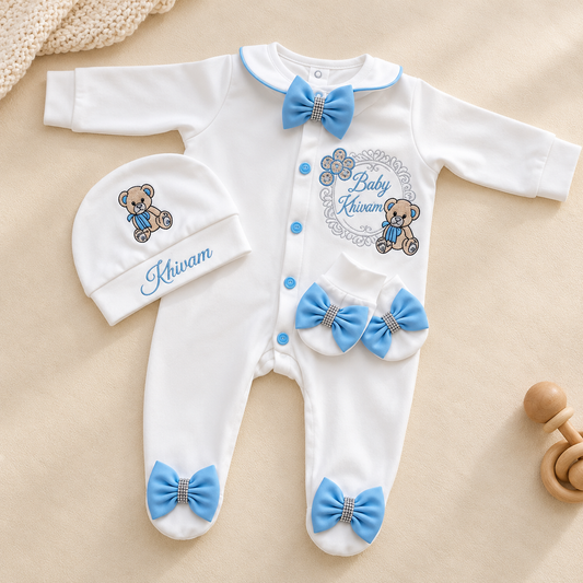 White long-sleeve baby footed sleeper with blue trim, blue bow details, and a teddy bear graphic reading 'Baby Kiwam,' shown with a matching beanie on a beige fabric background.