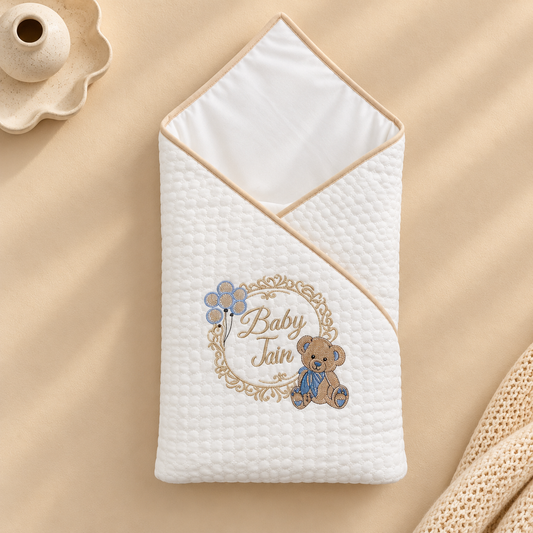 A white quilted baby swaddle wrap with a beige border, personalized with an embroidered teddy bear and the text 'Baby Jain' inside a decorative frame. The swaddle is styled on a beige fabric background with a small ceramic vessel and a knit fabric prop.