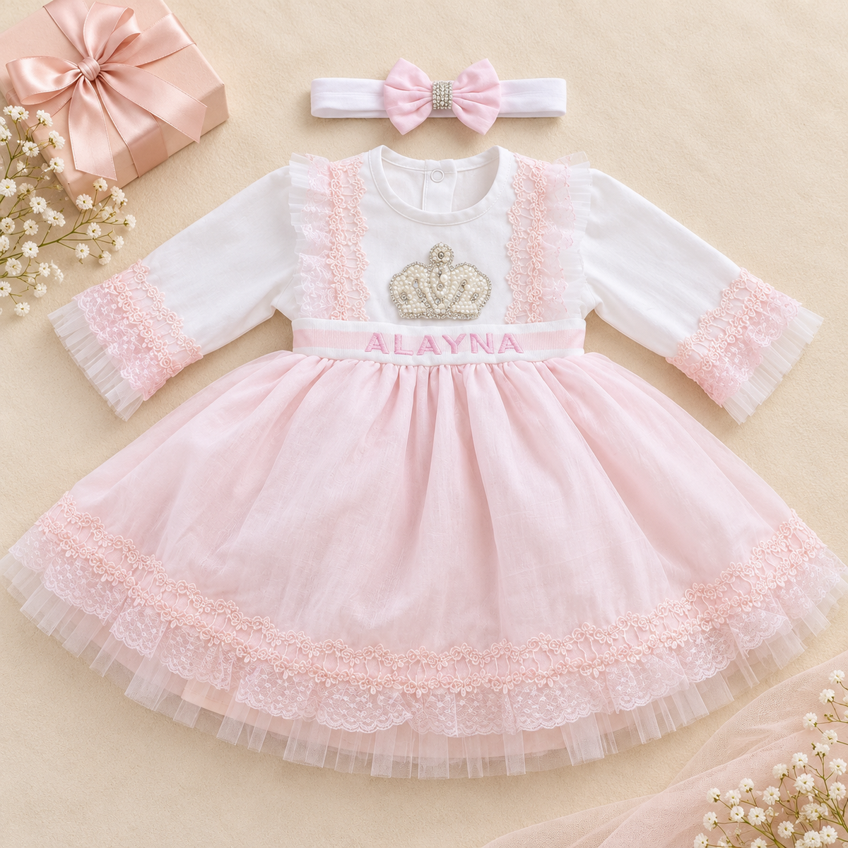 A baby girl's formal dress set featuring a white long-sleeve dress with a pink tulle skirt, lace trim, a pink name belt reading 'ALAYNA', and a matching pink bow headband, styled on a beige background with a gift box and baby flowers.
