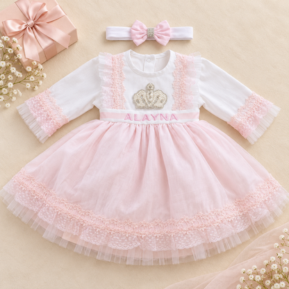 A baby girl's formal dress set featuring a white long-sleeve dress with a pink tulle skirt, lace trim, a pink name belt reading 'ALAYNA', and a matching pink bow headband, styled on a beige background with a gift box and baby flowers.