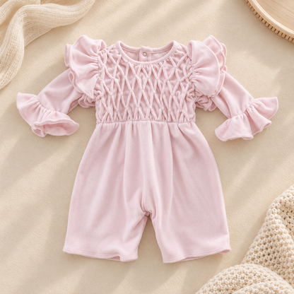A baby girl's pink long-sleeve romper with a smocked diamond-patterned bodice and ruffled sleeves, laid flat on a beige fabric background with styled knit props.