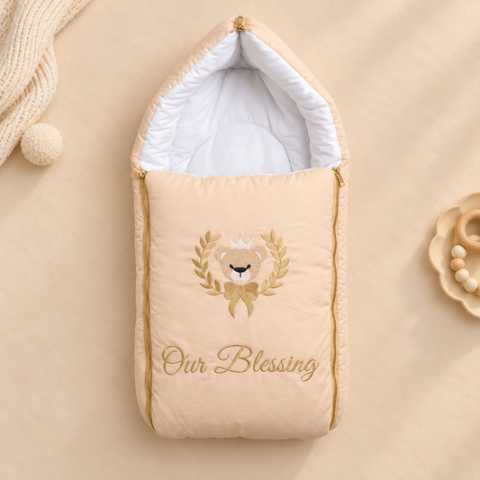 A beige baby footmuff with gold zipper trim and an embroidered teddy bear wreath with the words 'Our Blessing' on the front, shown on a matching beige background with soft styling props.