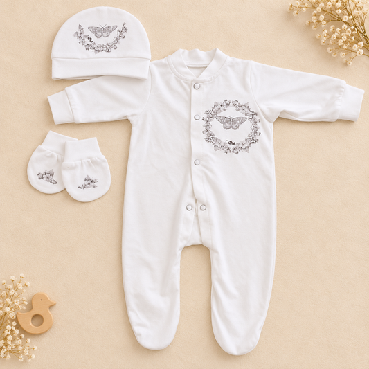 White baby footed sleeper set with a matching bonnet and mittens, laid flat on a beige background with decorative dried flowers and a wooden teether. The sleeper and bonnet have a gray embroidered winged motif inside a floral oval, and the mittens have a smaller matching motif.