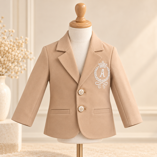 A beige children's blazer displayed on a white dress form mannequin, featuring a white embroidered monogram crest with the letter 'A' on the left chest. The blazer has two decorative gold-tone buttons and is set against a warm, softly lit background with a vase of dried flowers on the left.