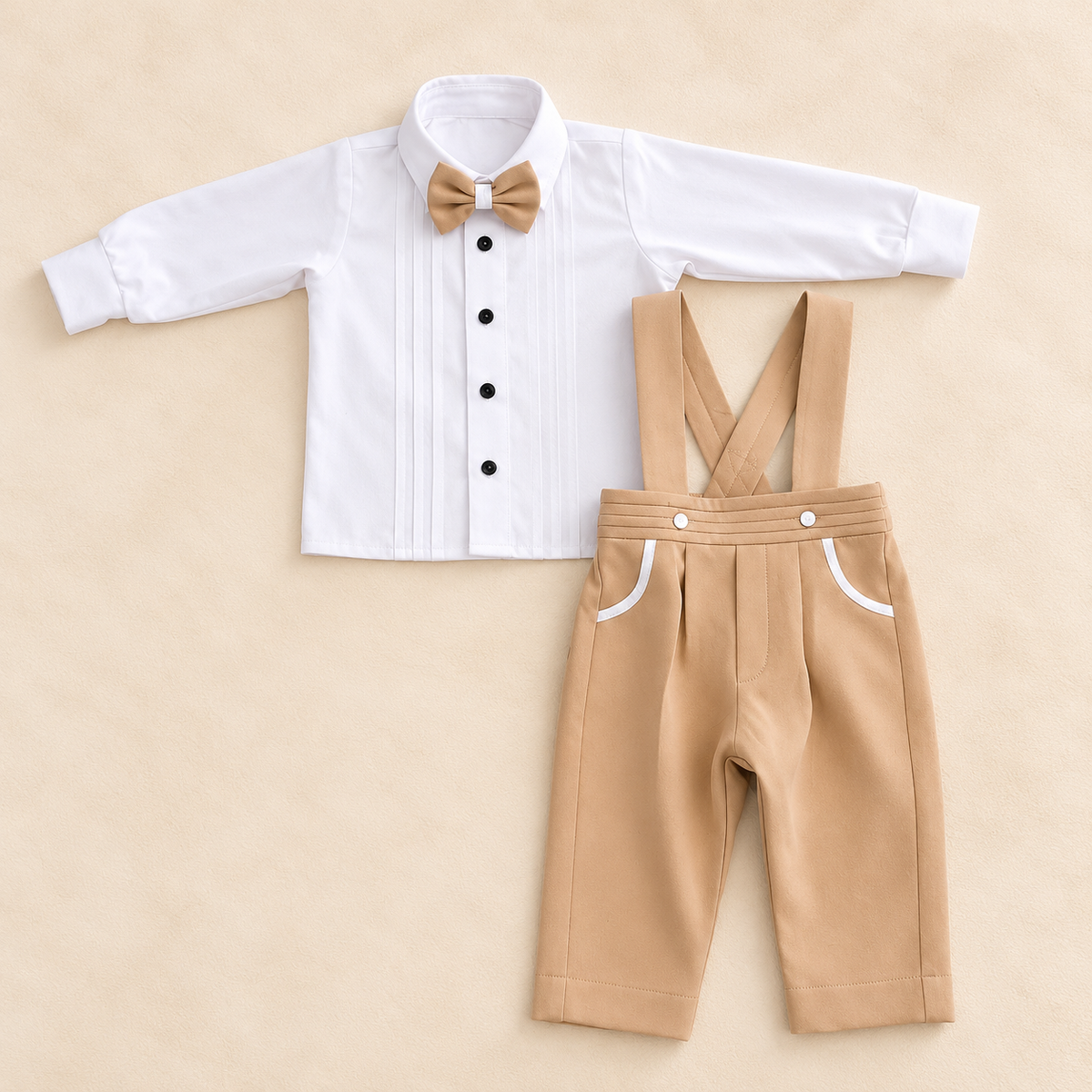 A baby boy's formal outfit set consisting of a white long-sleeve shirt with black buttons, a tan bow tie, and tan suspenders with white pocket trim, laid flat on a beige background.