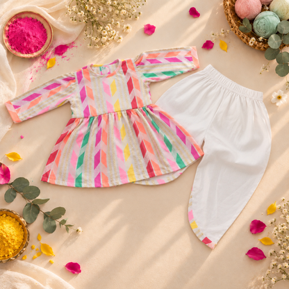 A baby girl's festive outfit featuring a long-sleeve peplum top with a colorful diagonal striped pattern in pink, orange, yellow, green, and blue on a white base, paired with solid white pants, styled in a Holi-themed flat lay with pink powder, flowers, and decorative props.