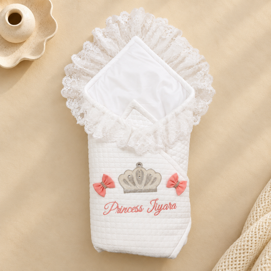 A white quilted baby swaddle blanket with a lace-trimmed hood, decorated with a silver crown, pink bows, and the embroidered text 'Princess Jyala' in pink. The swaddle is displayed flat on a beige fabric background with a decorative ceramic dish in the top-left corner.