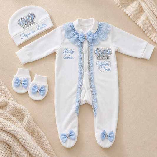 A baby gift set laid flat on a beige fabric background, including a white footless one-piece outfit with light blue lace trim, bow details, and embroidered text, a matching beanie with a crown graphic and text, and a pair of booties with light blue bows. Soft knit fabric props are visible around the items.
