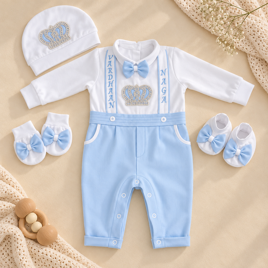 A baby boy's outfit set laid flat on a beige fabric background, including a white long-sleeve romper with light blue suspenders and bow tie, a light blue pair of pants, a matching white and blue bonnet, and white booties with blue bows. The romper has a crown graphic and the names 'VARDHAAN' and 'NAGI' printed vertically.