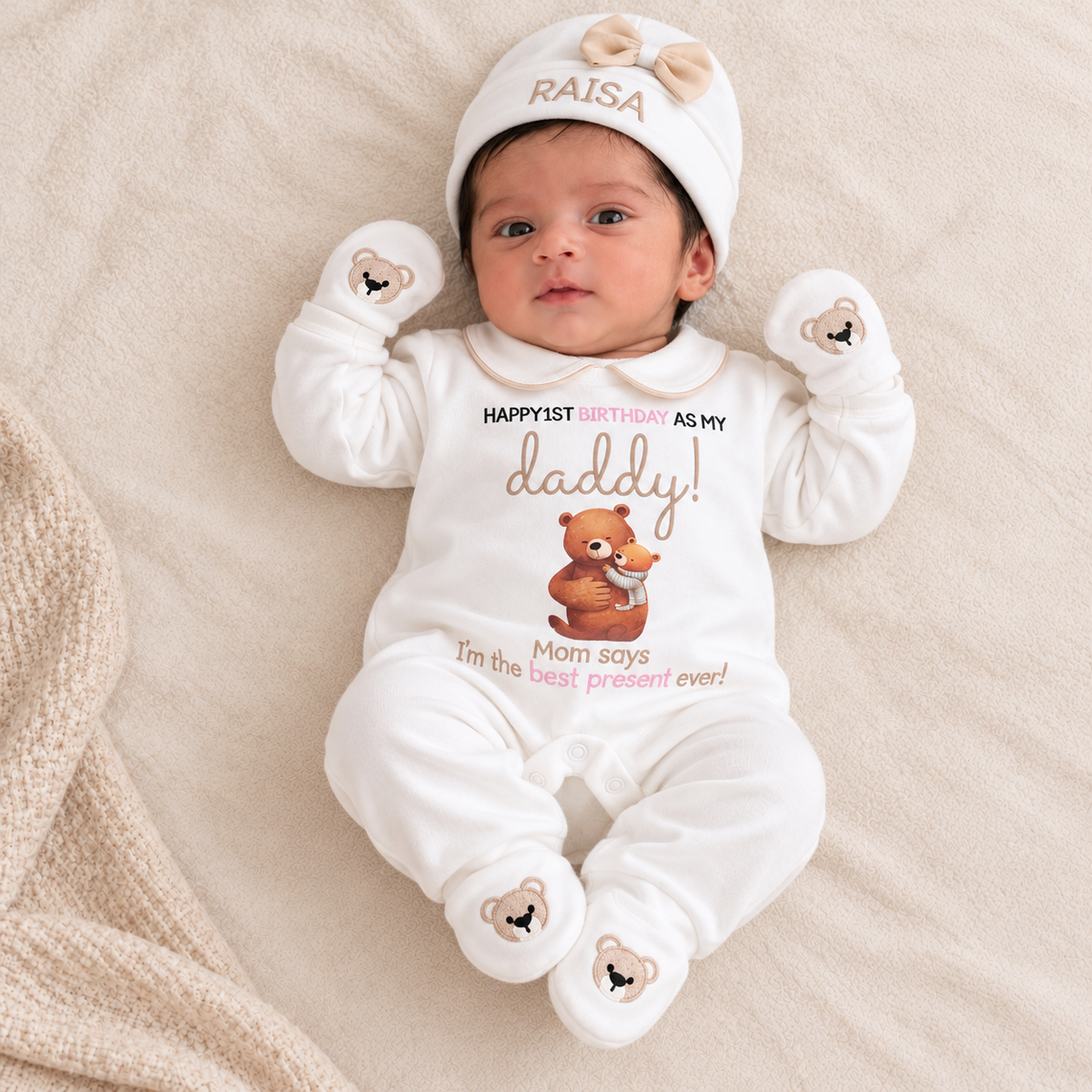A baby lying on a beige blanket wearing a white personalized onesie with a teddy bear graphic and the text 'HAPPYFIRST BIRTHDAY AS MY daddy! Mom says I'm the best present ever!', along with a matching white hat and mittens.