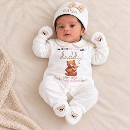 A baby lying on a beige blanket wearing a white personalized onesie with a teddy bear graphic and the text 'HAPPYFIRST BIRTHDAY AS MY daddy! Mom says I'm the best present ever!', along with a matching white hat and mittens.