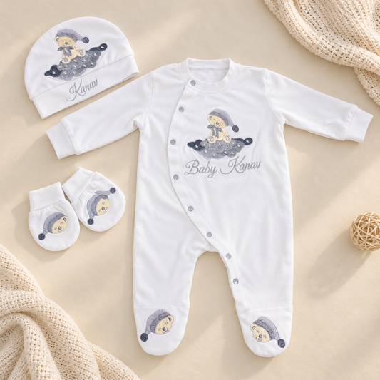 A white baby footed sleeper set with a matching beanie and mittens, featuring embroidered cartoon baby illustrations and the personalized name 'Baby Kanav' on a beige background with soft knit textiles.