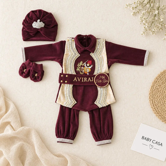A baby boy's traditional Indian outfit laid flat on a beige fabric background. The outfit consists of a deep maroon velvet long-sleeve tunic with cream and gold embroidered details, matching maroon pants, a maroon turban hat with a silver rhinestone bow, and maroon booties.